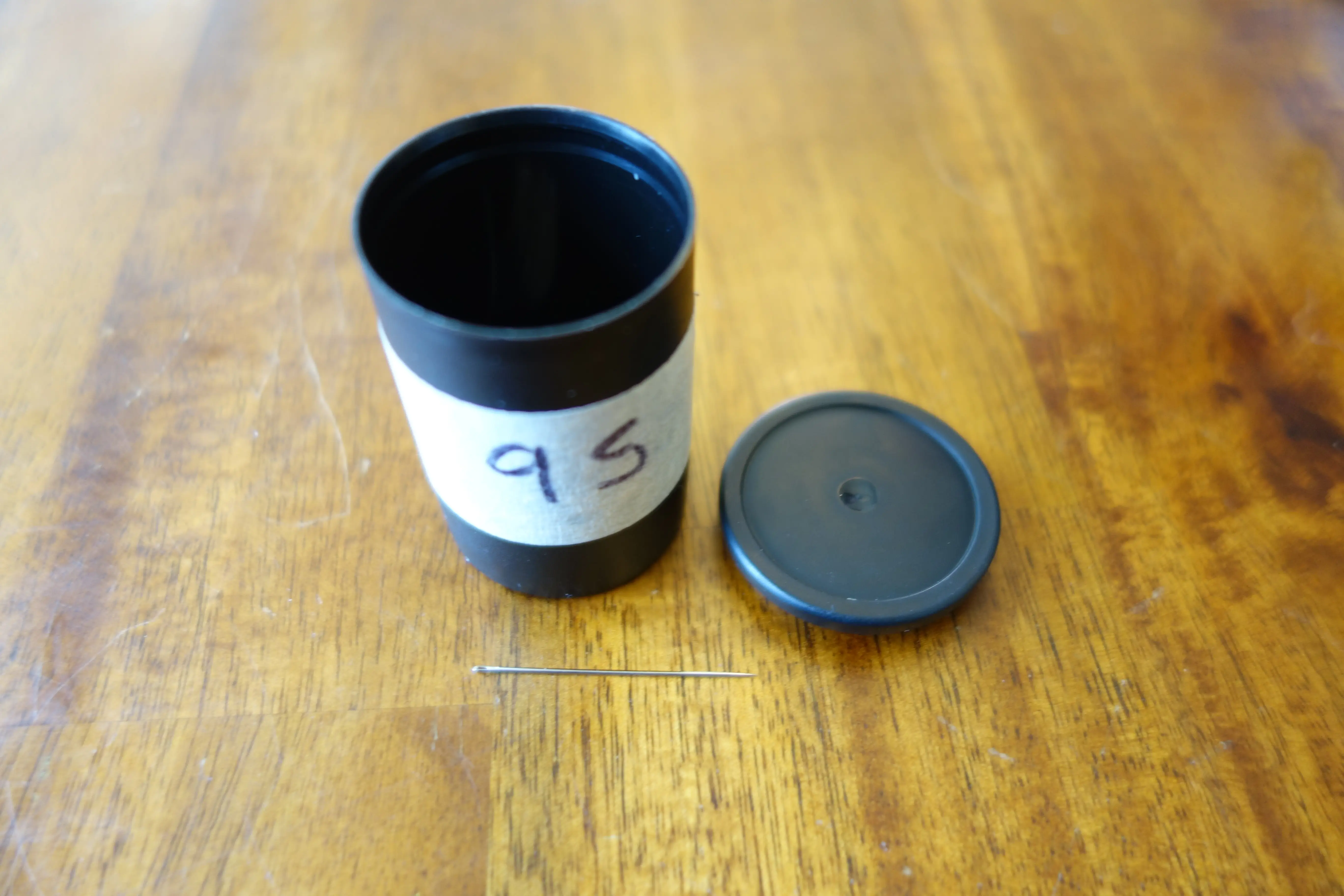 a black plastic film canister labeled '9 S' with the cap off and a needle sitting in front